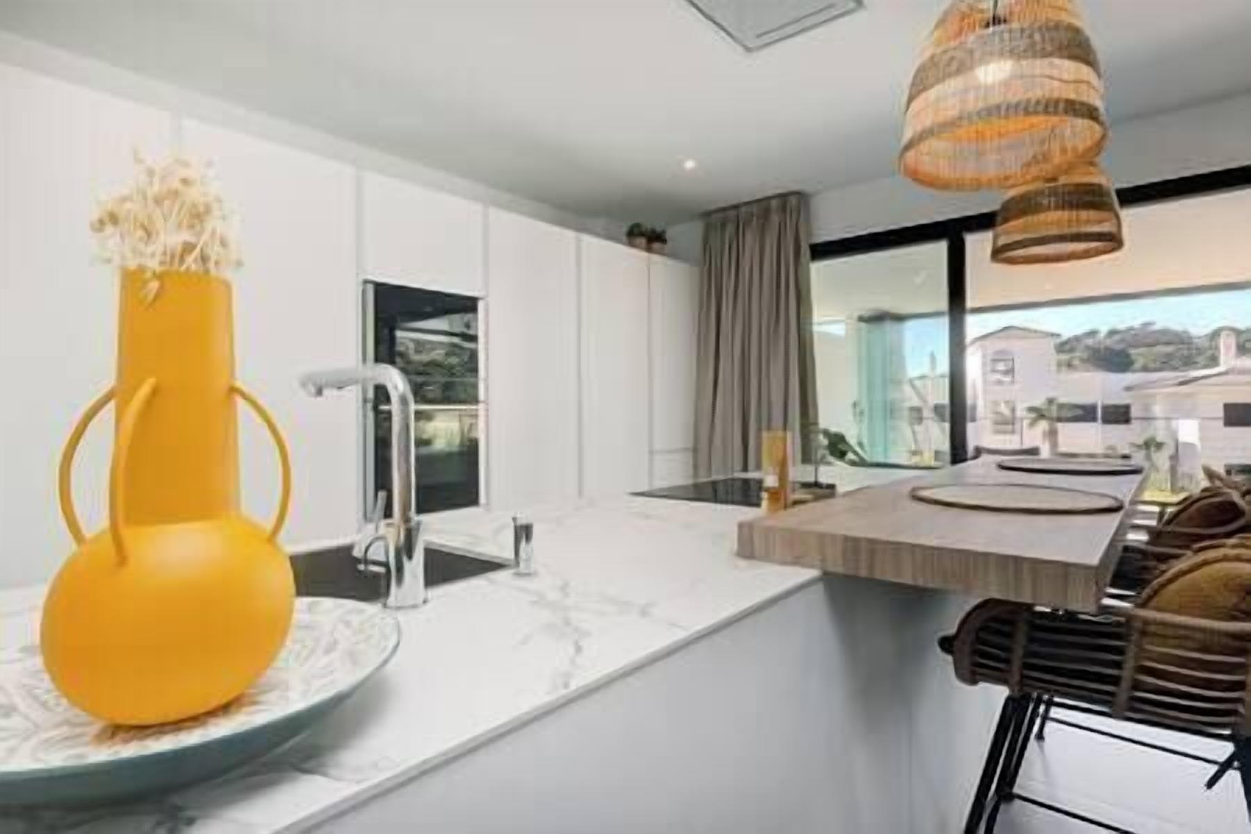 Een moderne keuken met een wit aanrecht, een gele vaas, en een eethoek met een houten tafel en stoelen.