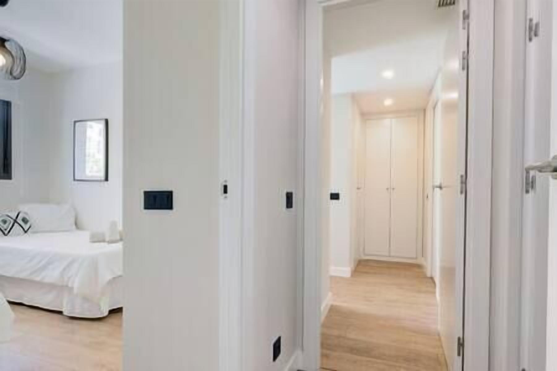 A hotel room with a bed, a bedside table, and a small window. There is a hallway leading to another room with a door.