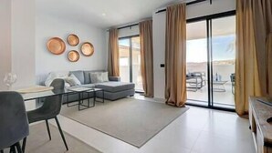 A modern living room with a sofa, a coffee table, and a dining area with chairs. There are large windows with curtains, and a view of the outdoors. - Luxurious Apartment @ Estepona Hills with views! (Estepona)