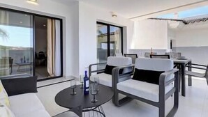 A modern outdoor seating area with two white cushioned chairs and a round table, featuring a glass partition and a view of the interior. - Luxurious Apartment @ Estepona Hills with views! (Estepona)