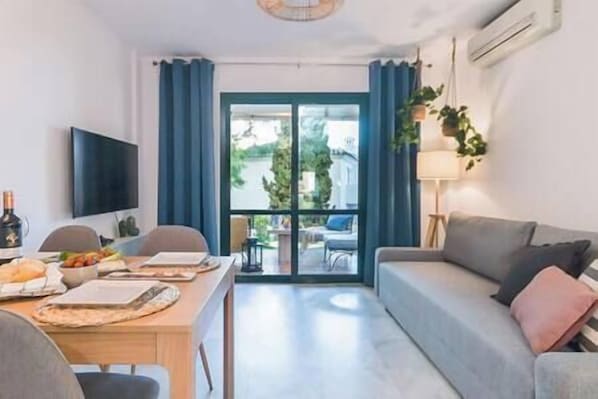 A modern living room with a sofa, a dining table with food, a TV, and a view of greenery outside.