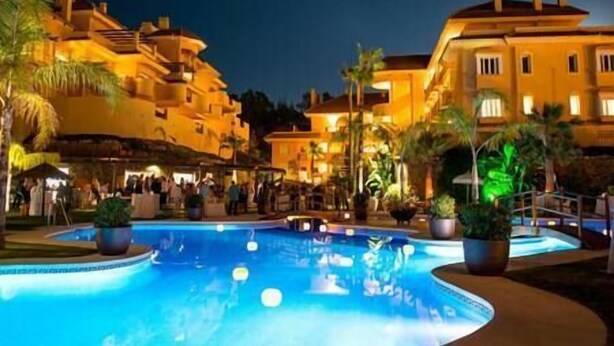 Piscina rodeada por palmeiras e edifícios com iluminação quente.
