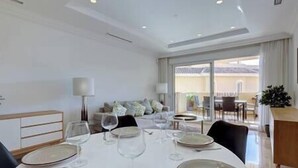 Uma sala de jantar com uma mesa branca posta para quatro pessoas, um sofá ao fundo e vista para o exterior através de grandes janelas.