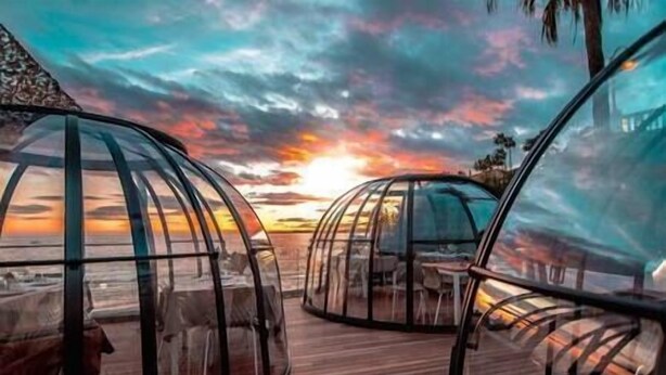 A transparent dome structure with a wooden deck, offering a view of the sunset.