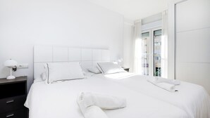 A modern hotel room with a large bed, white bedding, a nightstand with a lamp, and a view of a building through a glass door.