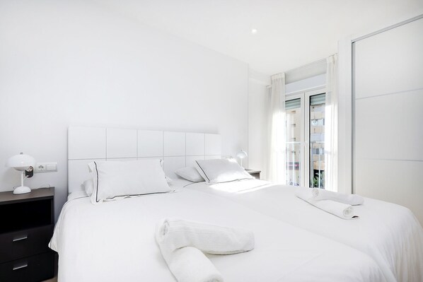 A modern hotel room with a large bed, white bedding, a nightstand with a lamp, and a view of a building through a glass door. - Stunning 1 bedroom apartment in Marbella Centre (Marbella)