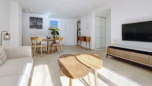 A modern living room with a sofa, a TV, a dining area with chairs and a table, and a kitchen in the background.