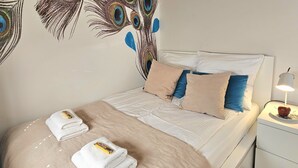 A neatly made bed with a peacock feather wall art, a bedside lamp, and a stack of towels. - Apartament Pawie Oczko - 365PAM (Kolobrzeg)