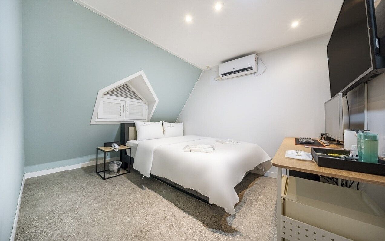 A bedroom with a bed, a desk, an air conditioner, a telephone, and a trash bin.