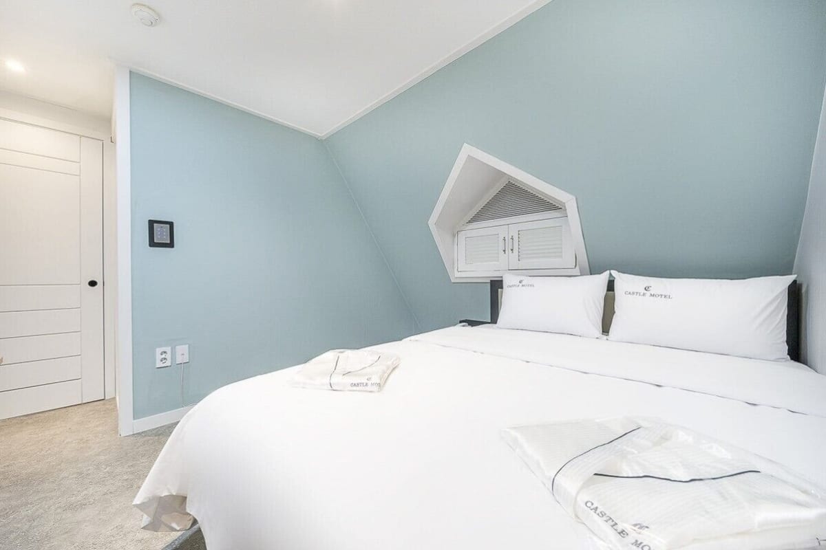 Un dormitorio con una cama, una puerta blanca, un ventanal pequeño y un interruptor de luz en la pared.