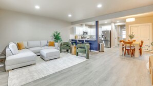 Un salon moderne avec un canapé d’angle, un coin repas avec une table et des chaises en bois, et une cuisine en arrière-plan.