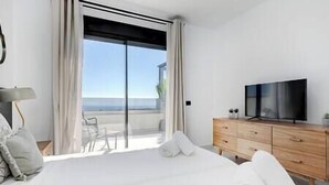 A modern hotel room with a large bed, a TV, a wooden dresser, and a balcony with a view of the sea. - New 2 Bed Penthouse @ Estepona Hills With Sea View (Estepona)