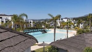 A resort with a swimming pool, palm trees, and thatched-roof structures. - New 2 Bed Penthouse @ Estepona Hills With Sea View (Estepona)