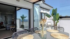 An outdoor dining area with a table set for four, surrounded by chairs. There is a glass partition to the left, and a door leading to an interior space. - New 2 Bed Penthouse @ Estepona Hills With Sea View (Estepona)