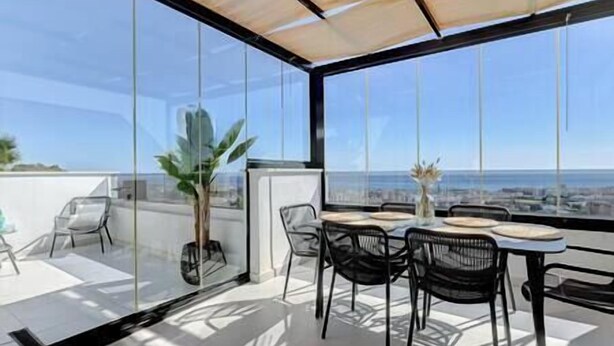 A modern outdoor dining area with a glass enclosure, a table set with plates and a vase, and a view of the ocean. - New 2 Bed Penthouse @ Estepona Hills With Sea View (Estepona)