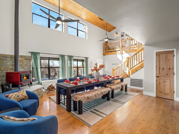 Un salon spacieux avec une cheminée, un coin repas agrémenté d’un escalier en bois et de grandes fenêtres.