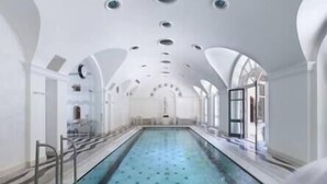 Une longue et étroite piscine intérieure, avec une eau d’un bleu limpide, entourée de murs blancs et surmontée de voûtes.