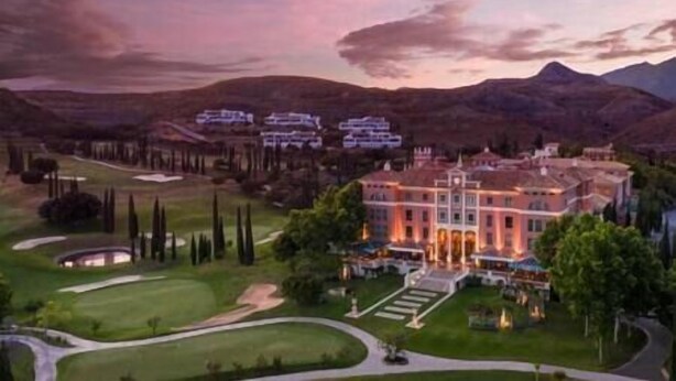 Un grand hôtel, bien éclairé, avec une entrée imposante, entouré de jardins soignés et d’un parcours de golf.
