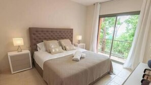 Une chambre d’hôtel avec un grand lit, deux tables de chevet avec des lampes, une vue sur la verdure par la fenêtre et un balcon.