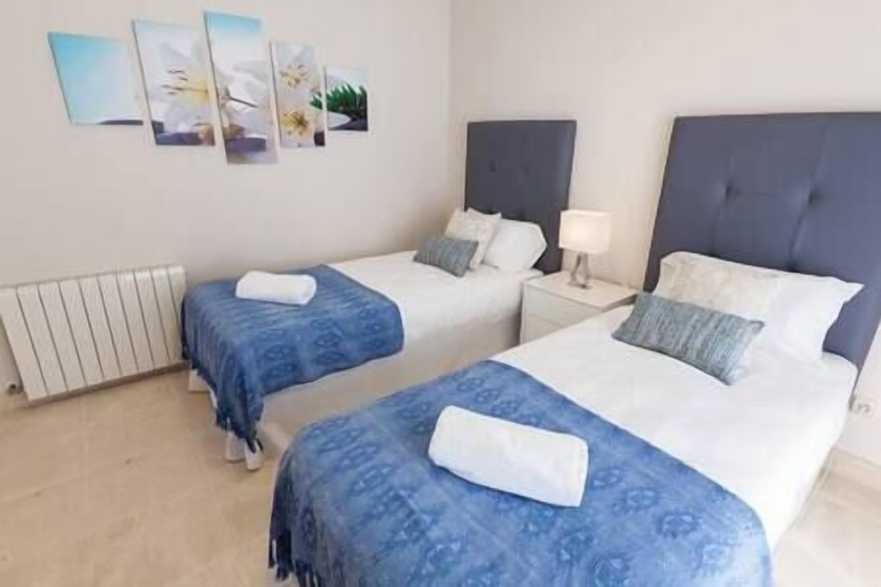 Ein Hotelzimmer mit zwei Betten, jeweils mit blauer Bettdecke und weißen Kissen. Darüber hängen Bilder an der Wand und links befindet sich ein weißer Heizkörper.
