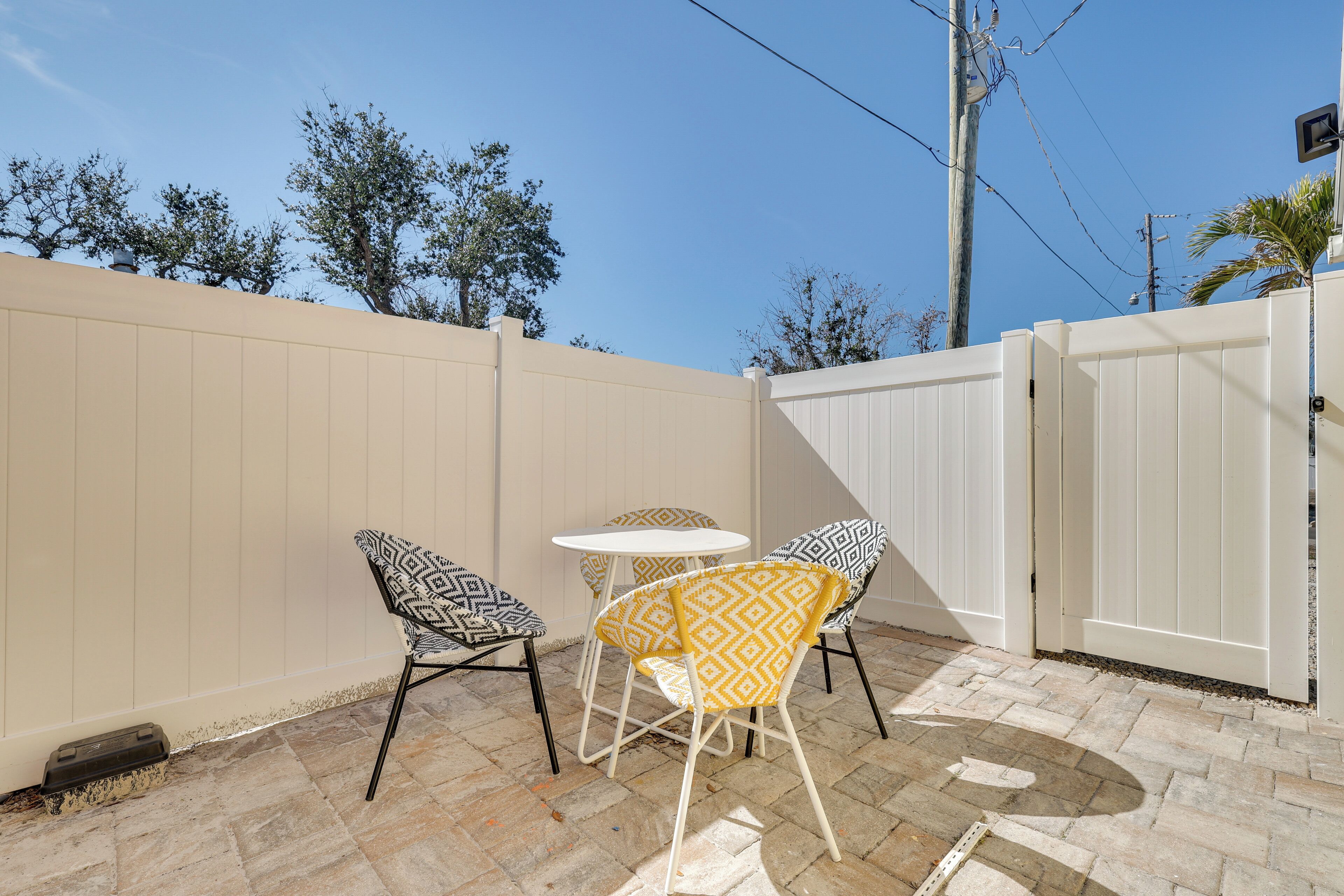 An outdoor patio area with a white table and two patterned chairs, a beige fence, and a palm tree in the background.