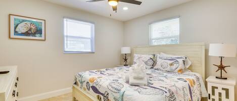 A bedroom with a bed, ceiling fan, two windows, and a nightstand with a lamp.