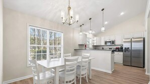 A modern kitchen with white cabinetry, stainless steel appliances, and a dining area with white chairs and a table. - Oceanside Village - Beautiful 3BR 2BA - Golf Cart Included! (Surfside Beach)