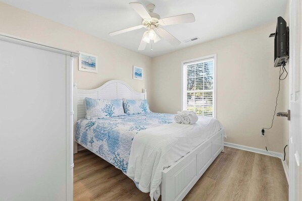 A bedroom with a bed, a ceiling fan, a window with blinds, and a wall-mounted TV.