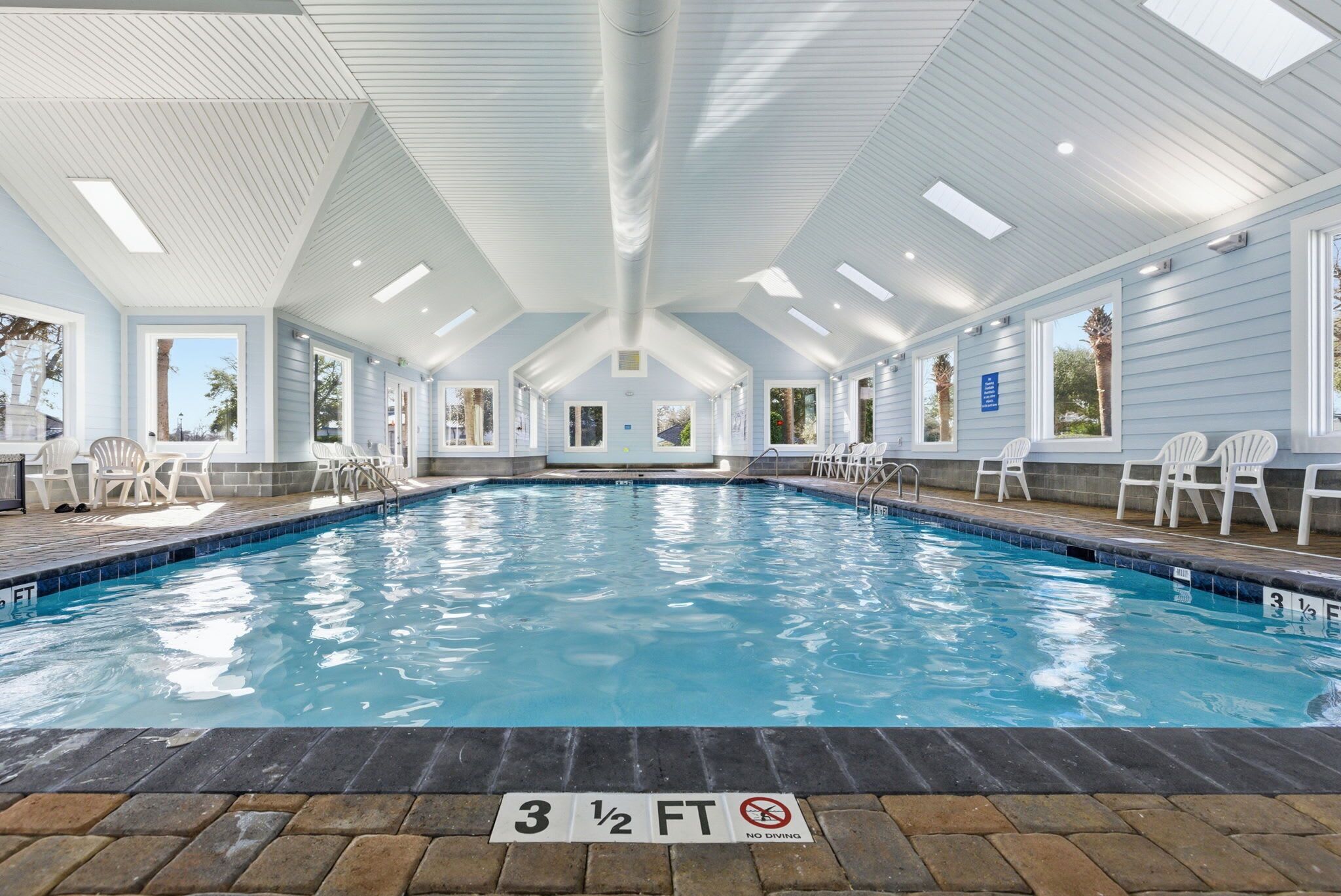 An indoor swimming pool with a 3 1/2 ft depth, surrounded by white chairs and a covered roof with skylights.