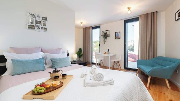 Ein modernes Hotelzimmer mit einem großen Bett, einem Frühstückswagen, einem blauen Sessel und Blick ins Freie.