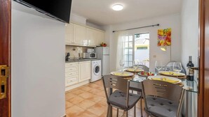 Een kleine keuken met witte kastjes, een magnetron en een wasmachine. Er is een eettafel met stoelen en een fles wijn.