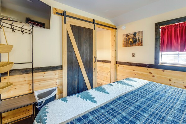 Une chambre à coucher avec un lit, une tête de lit en bois, une porte coulissante, un téléviseur fixé au mur, une fenêtre avec un rideau rouge et un porte-manteau mural.