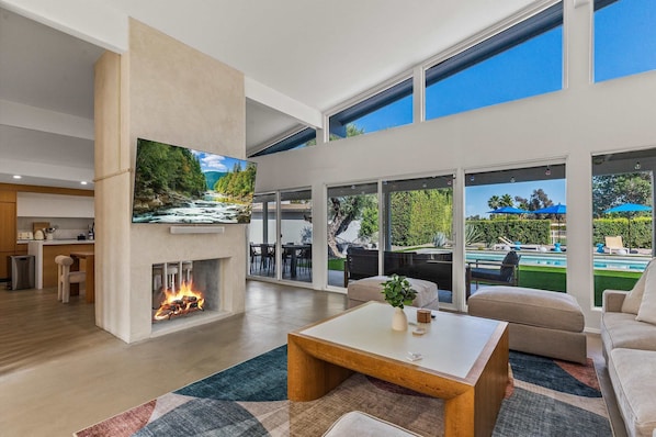 A modern living room with a fireplace, a flat-screen TV, and a view of a pool and outdoor seating.