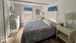 A bedroom with a bed, a dresser, a vanity with a mirror, a window with blue curtains, and a bedside table with a lamp. - The perfect cozy Chatham getaway awaits you! (Chatham)