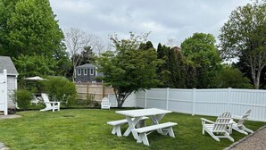 A backyard with a picnic table and Adirondack chairs, surrounded by a white fence and lush greenery. - The perfect cozy Chatham getaway awaits you! (Chatham)