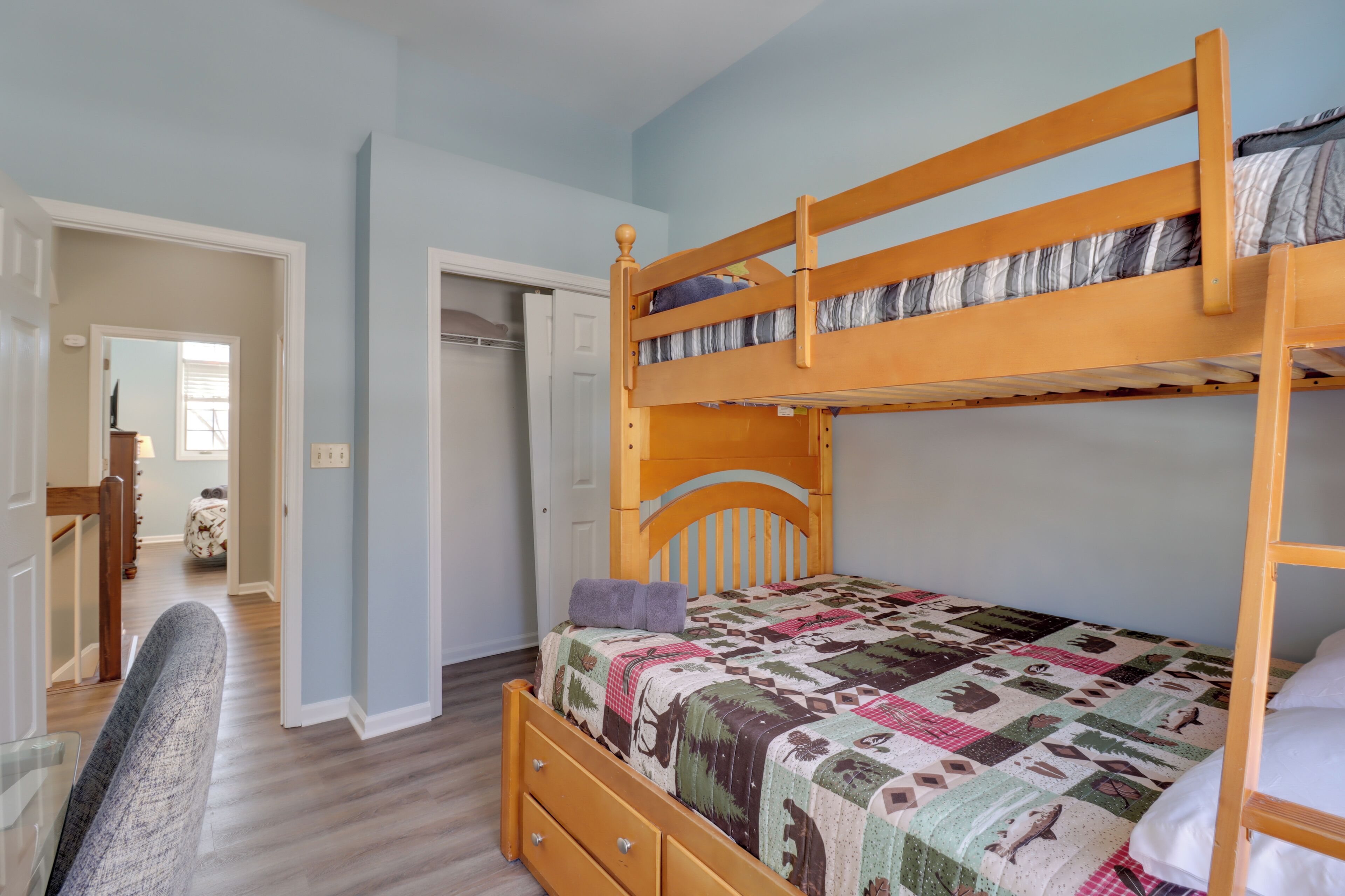 Ein Stockbett mit einer Tagesdecke, ein Holztisch und ein Stuhl in einem Zimmer mit hellblauen Wänden und Holzboden.