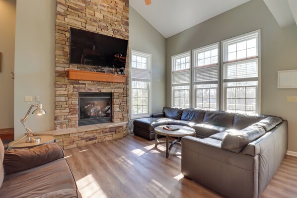 A living room with a stone fireplace, a leather sofa, a coffee table, and large windows.