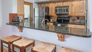 A kitchen with wooden cabinets, stainless steel appliances, and a granite countertop. There are three woven stools at the counter.