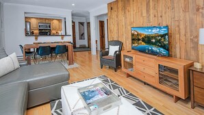 A modern living room with a grey sofa, a coffee table with magazines, a wooden TV stand, and a large TV displaying a scenic landscape.