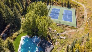 An aerial view of a property with a tennis court and a swimming pool surrounded by dense forest.