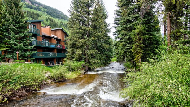 A multi-story building with balconies overlooking a flowing river, surrounded by dense forest.
