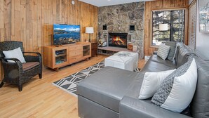 A living room with a stone fireplace, wooden panel walls, a grey sofa, and a television.