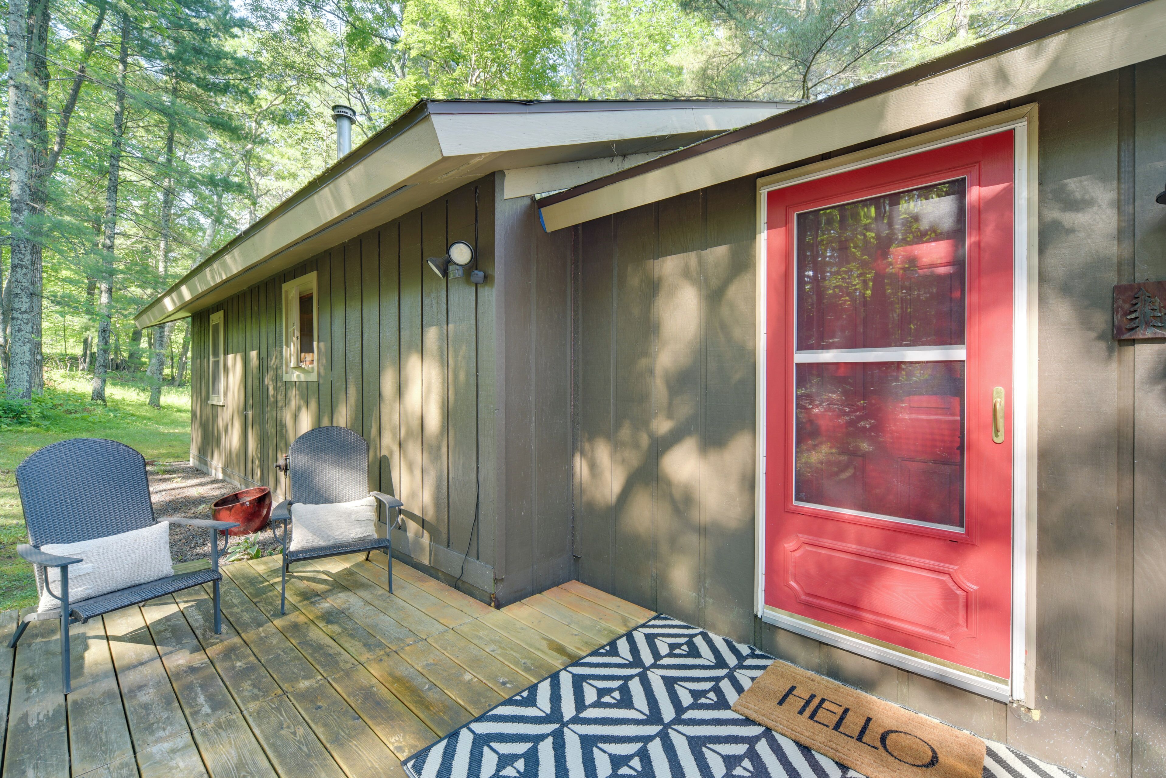 A wooden cabin with a red door, a porch with two chairs, and a welcome mat.