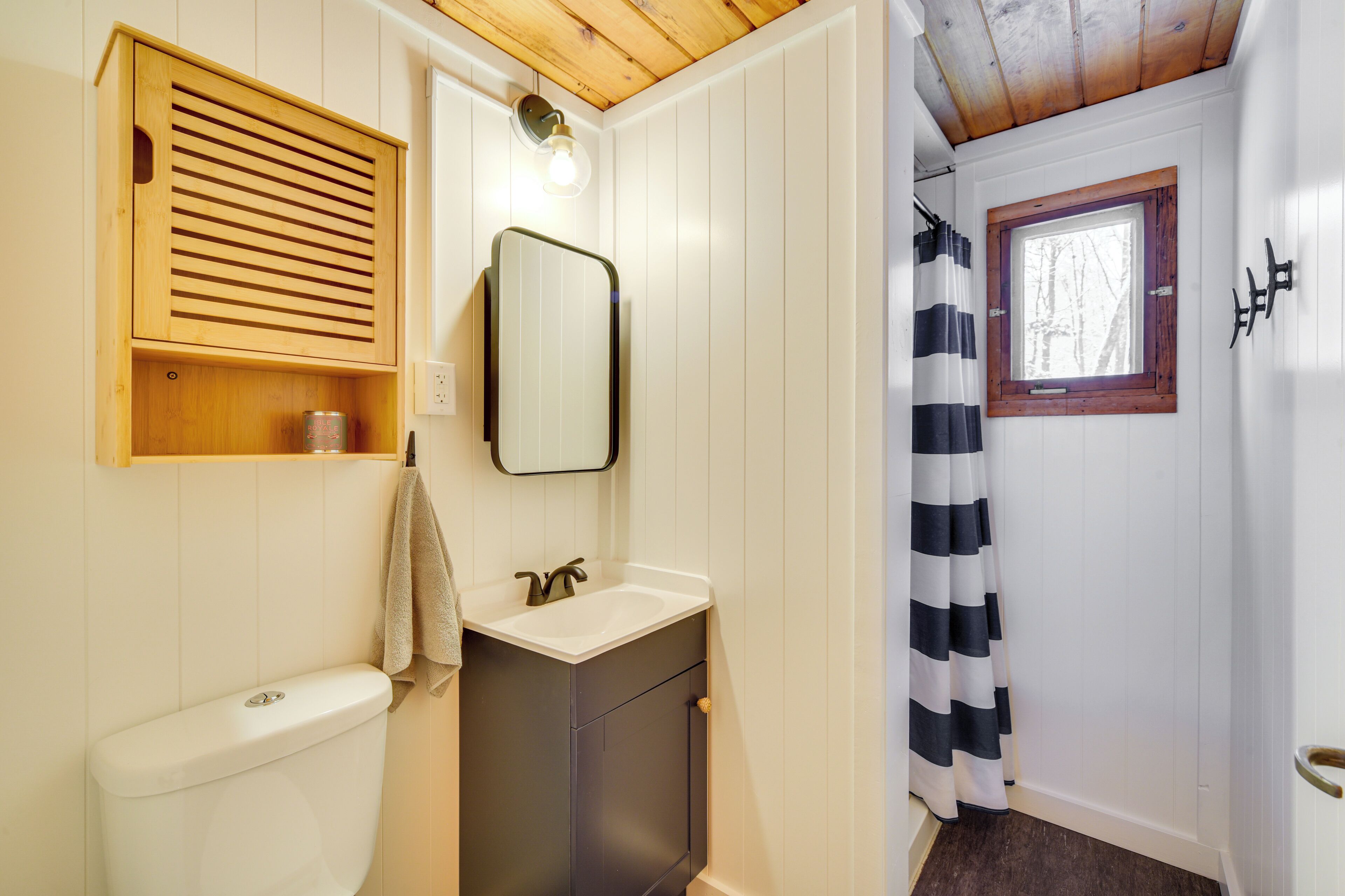 Ein Badezimmer mit Toilette, Waschbecken, Spiegel, Holzschrank und Fenster mit Holzrahmen.