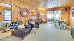 A cozy room with wooden walls and ceiling, a bed with a checkered blanket, a leather armchair, a small stool, a table with chairs, and a wicker basket on the wall. - Charming Cabin on Grindstone Lake (Hayward)