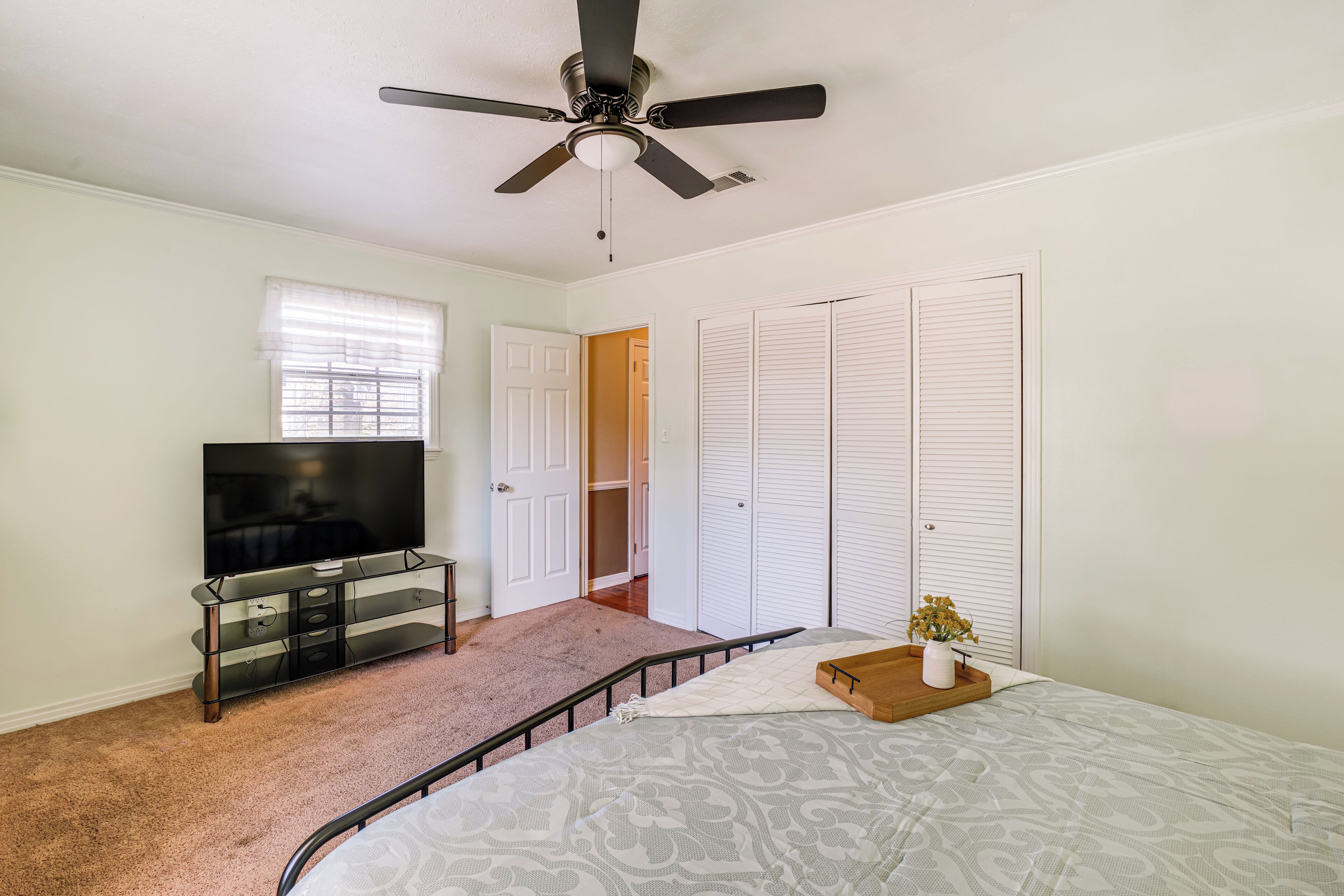 A bedroom with a bed, a TV on a glass cabinet, a ceiling fan, and a closet.