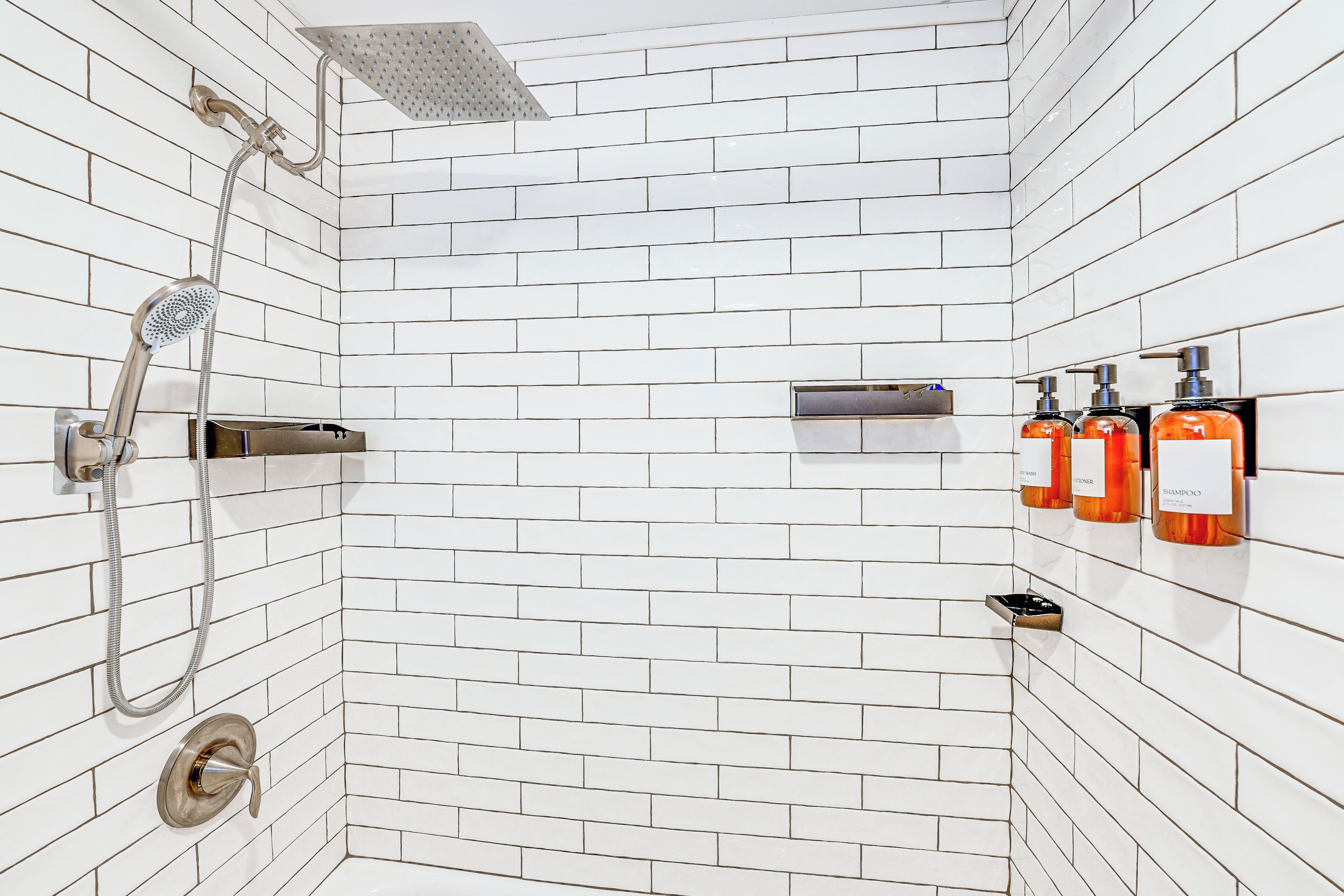 A bathroom with white subway tiles, a showerhead, a handheld shower, and three shampoo bottles on the wall.