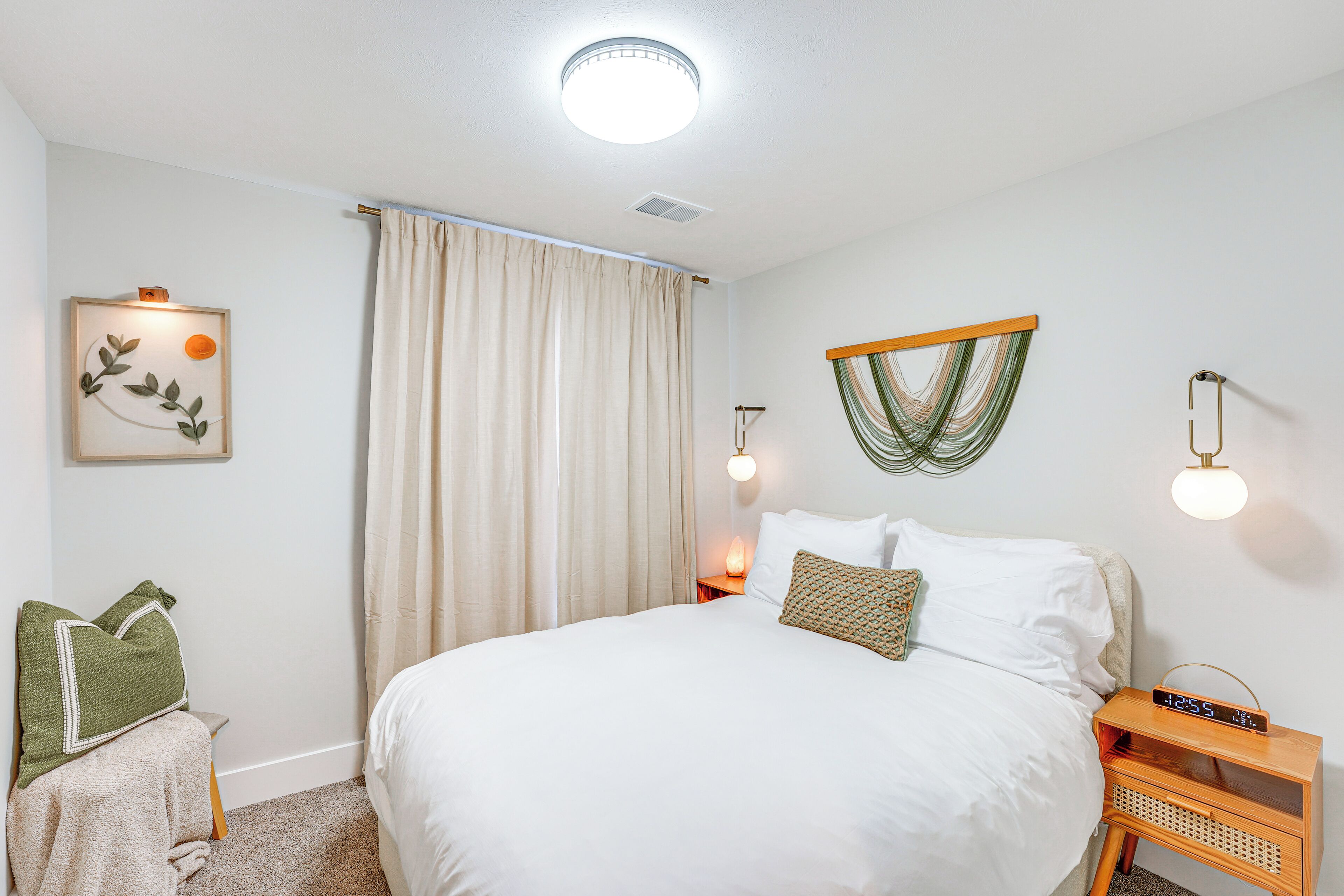 A neatly arranged bedroom with a large bed, a bedside table with a clock, a wall-mounted plant, and a window with curtains.