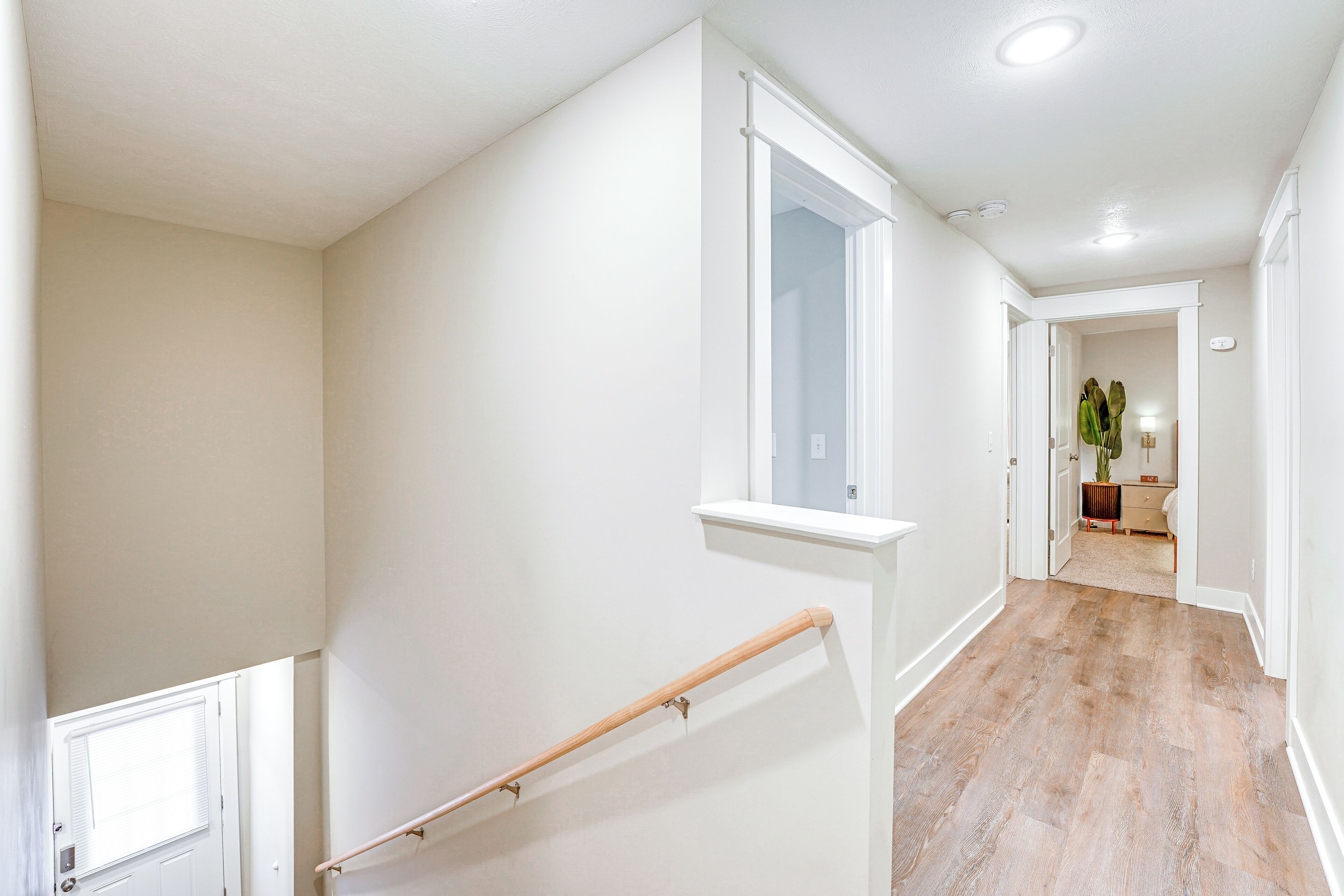 Un pasillo limpio y bien iluminado con piso de madera y paredes blancas, que conduce a una habitación con un ventanal grande y una planta.