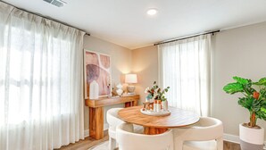 A dining area with a wooden table and white chairs, a lamp, a potted plant, and a painting on the wall.